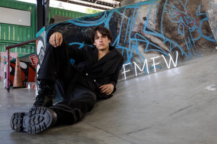 Young man in a black outfit sitting on a graffiti-covered skate ramp, leaning against the wall with 'FMW' neon letters nearby.